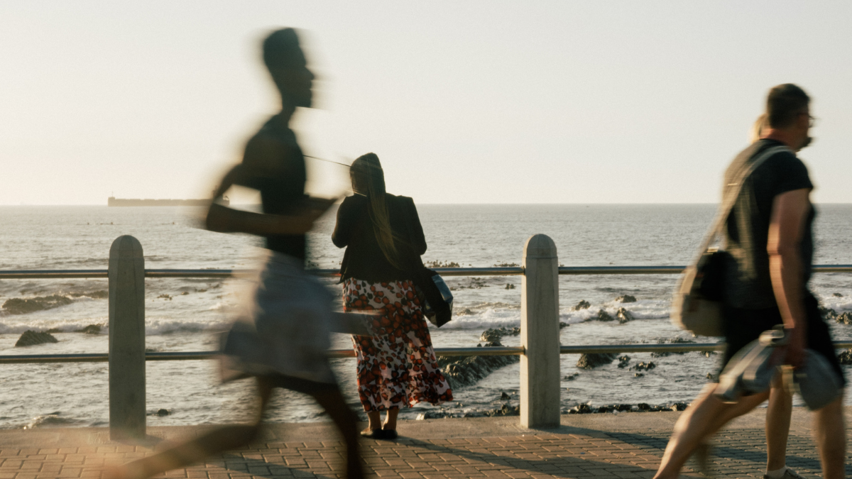 Person standing looking at the sea while runners and people walking are in the front of the photo 