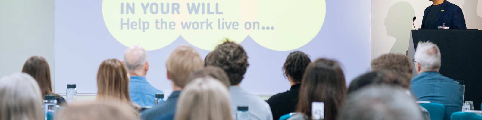 Image of a room full of delegates listening to a speaker and looking at a slide with the Remember a Charity logo on it