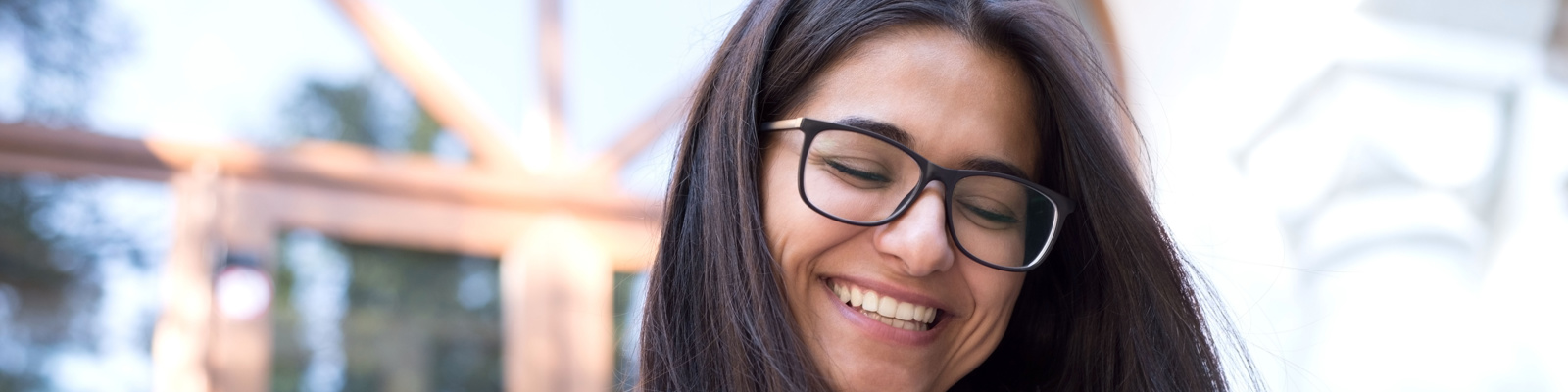Woman in glasses and a white shirt smiling whilst using a tablet outdoors with buildings blurred in the background