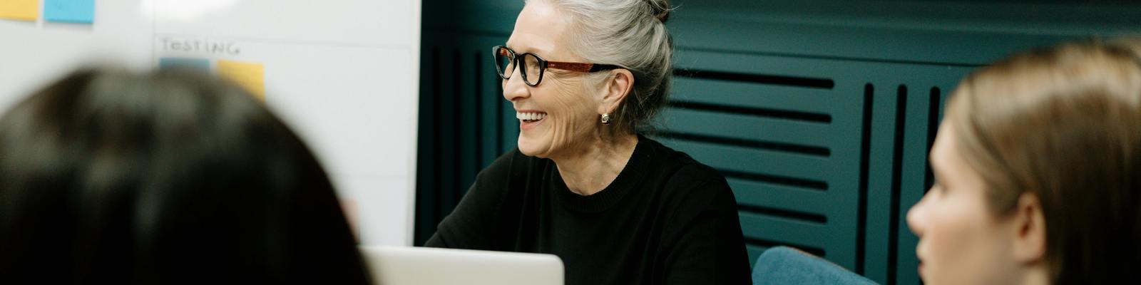 Older woman sat working on a laptop, leading a meeting. Two other professionals sit in the foreground out of focus. 