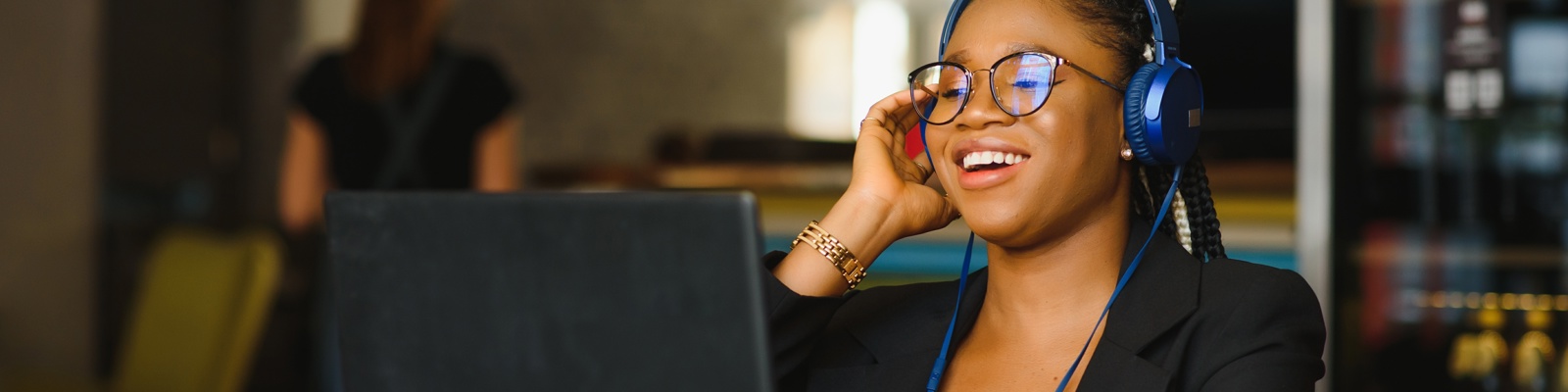 Photo of a young woman with glasses and a headset on talking and smiling whilst working on her tablet/laptop