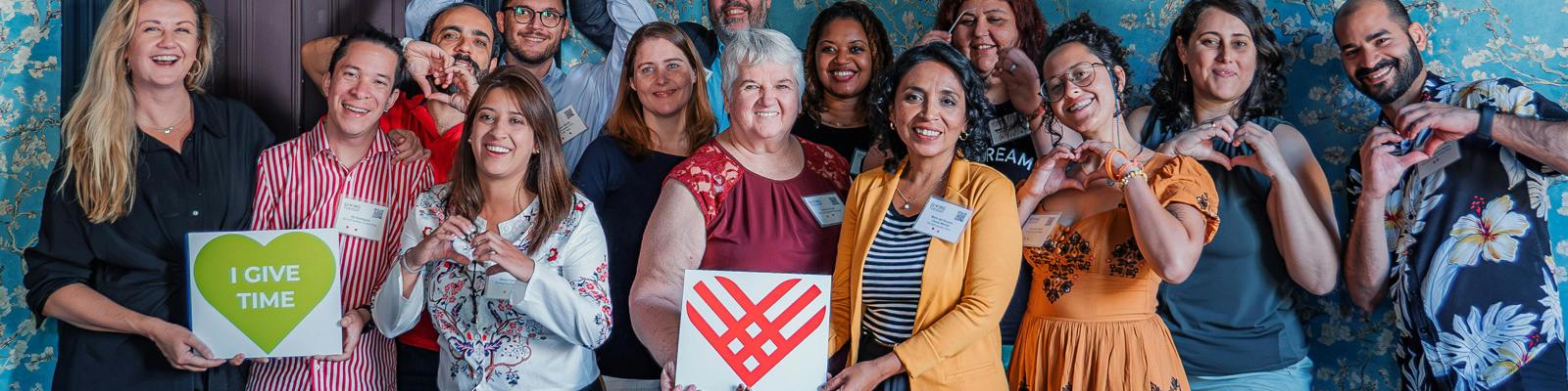 A photo of a group of people holding photos of the Giving Tuesday Logo, one man doing a heart shape with his hand, and one poster saying 'I Give Time'