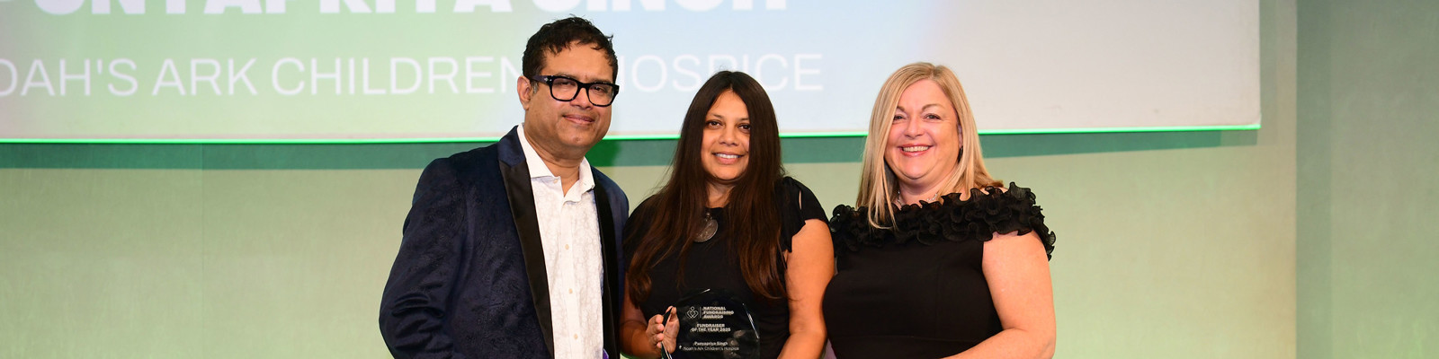 Photo of the Fundraiser Of The Year 2025 winner, Priya Singh, Being Handed Award by Awards Host, Paul Sinha, and Joanne Coe from Conference Care who Sponsored the Category.