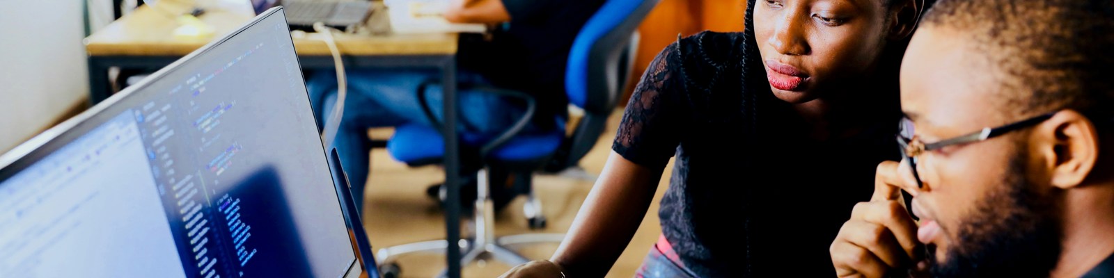 Two black people sit at a desk looking at a computer with data on the screen. 