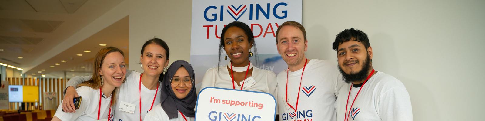A Group of People Wearing Giving Tuesdat T-Shirts. One in the Middle is Holding a Sign Up That Says 'I'm Supporting Giving Tuesday'. The Giving Tuesday Logo is in the Background on the Wall.